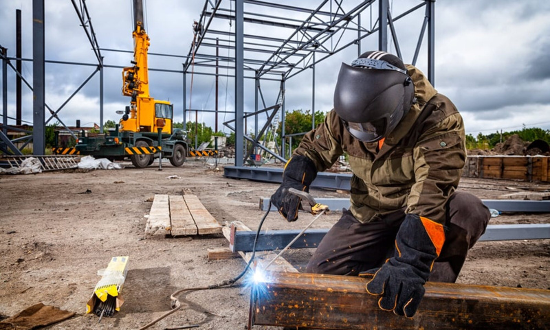Welder joining structural steel on a job site with a crane in the background