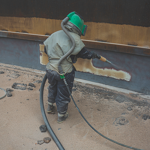 Operator blasting old coating off a large steel plate wall